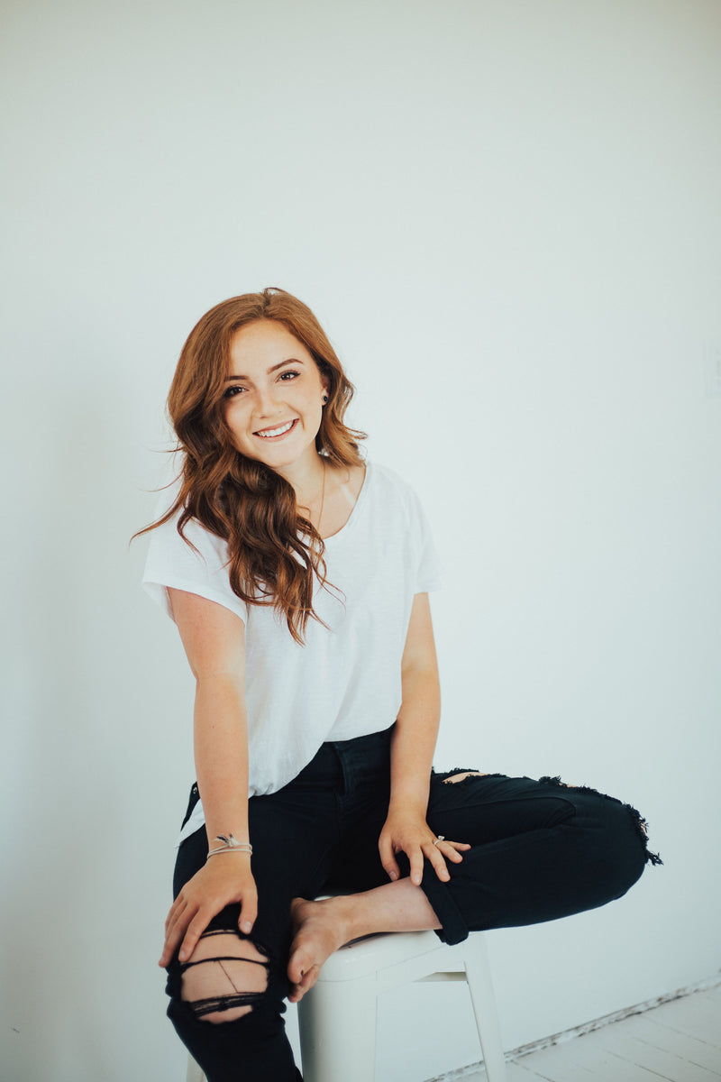 Portrait of a smiling woman with auburn hair seated on a stool in a bright studio